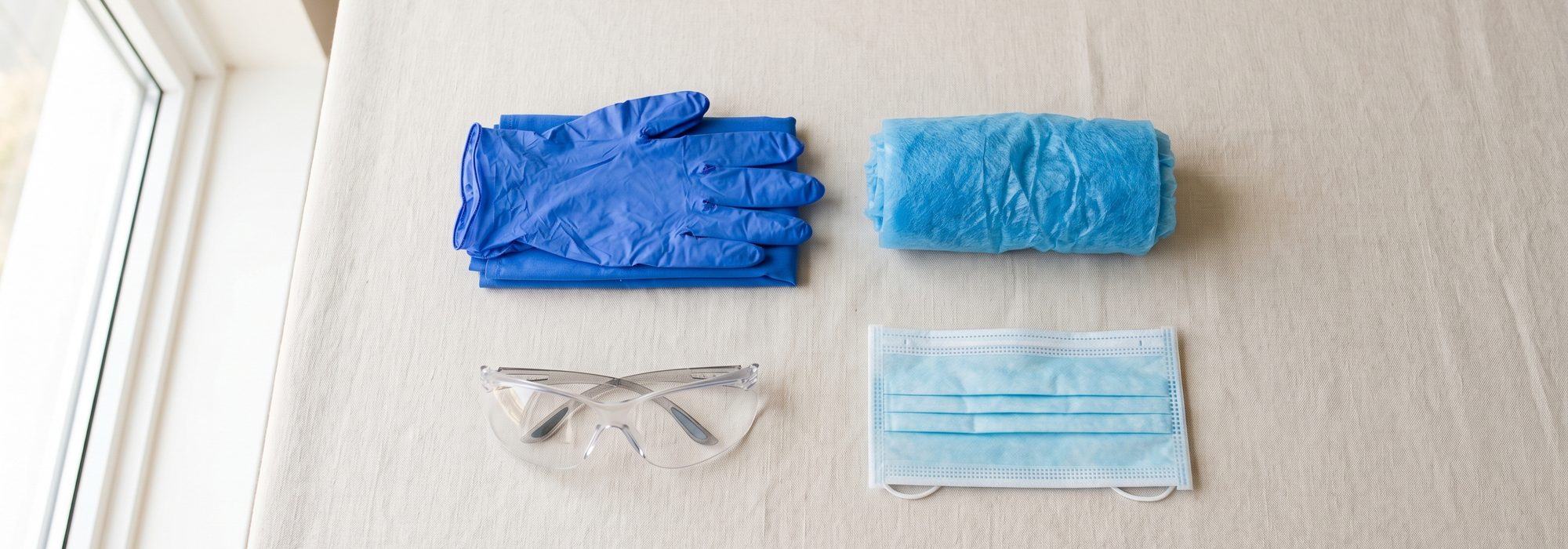 Flat-lay of blue nitrile gloves, bouffant cap, safety glasses and face mask
